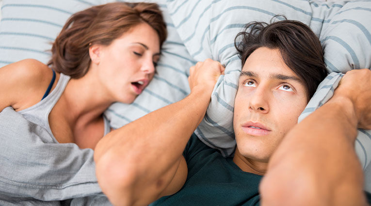 man covers ears with pillow as bed partner snores