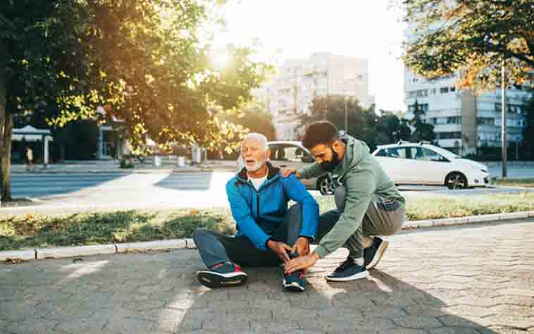 Adult son helps senior father upon falling, while he holds his ankle.