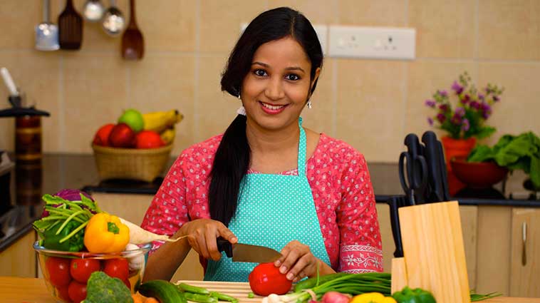 Woman wearing an apron and sitting at a table cutting fruits and vegetables.