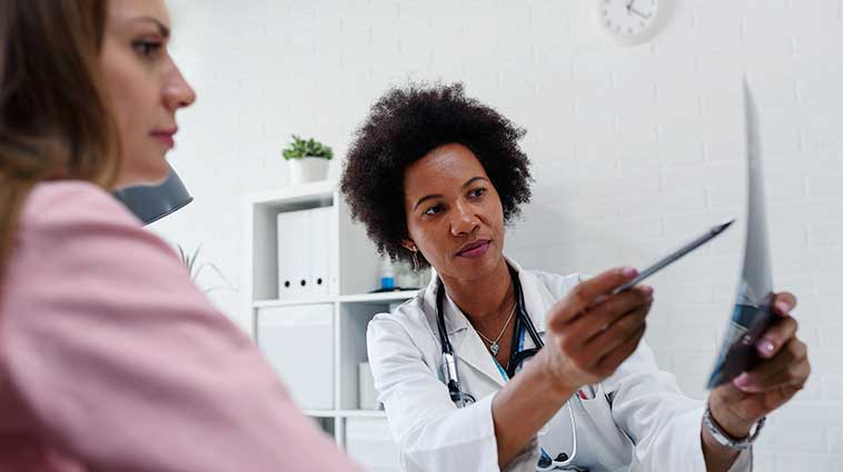 Black female physician wearing a white lab coat shares mammogram results with white female patient in a pink top.