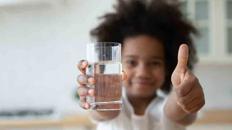 African Amercian child holding up a glass of water with a thumbs up for a blog about the health benefits of drinking water.