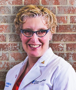Jennifer Manders, MD, breast surgeon, wearing a white lab coat and standing in front of a brick wall.