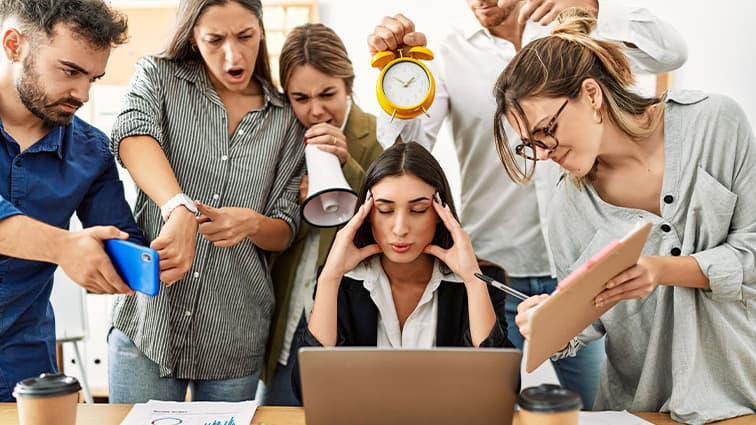 Person seated in front of laptop computer with hands on forehead. The person is surrounded by others who are causing stress.